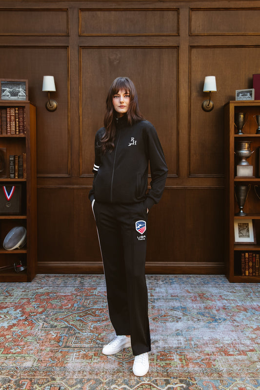 Person wearing a black tracksuit with logos in a room with wooden paneling and bookshelves.