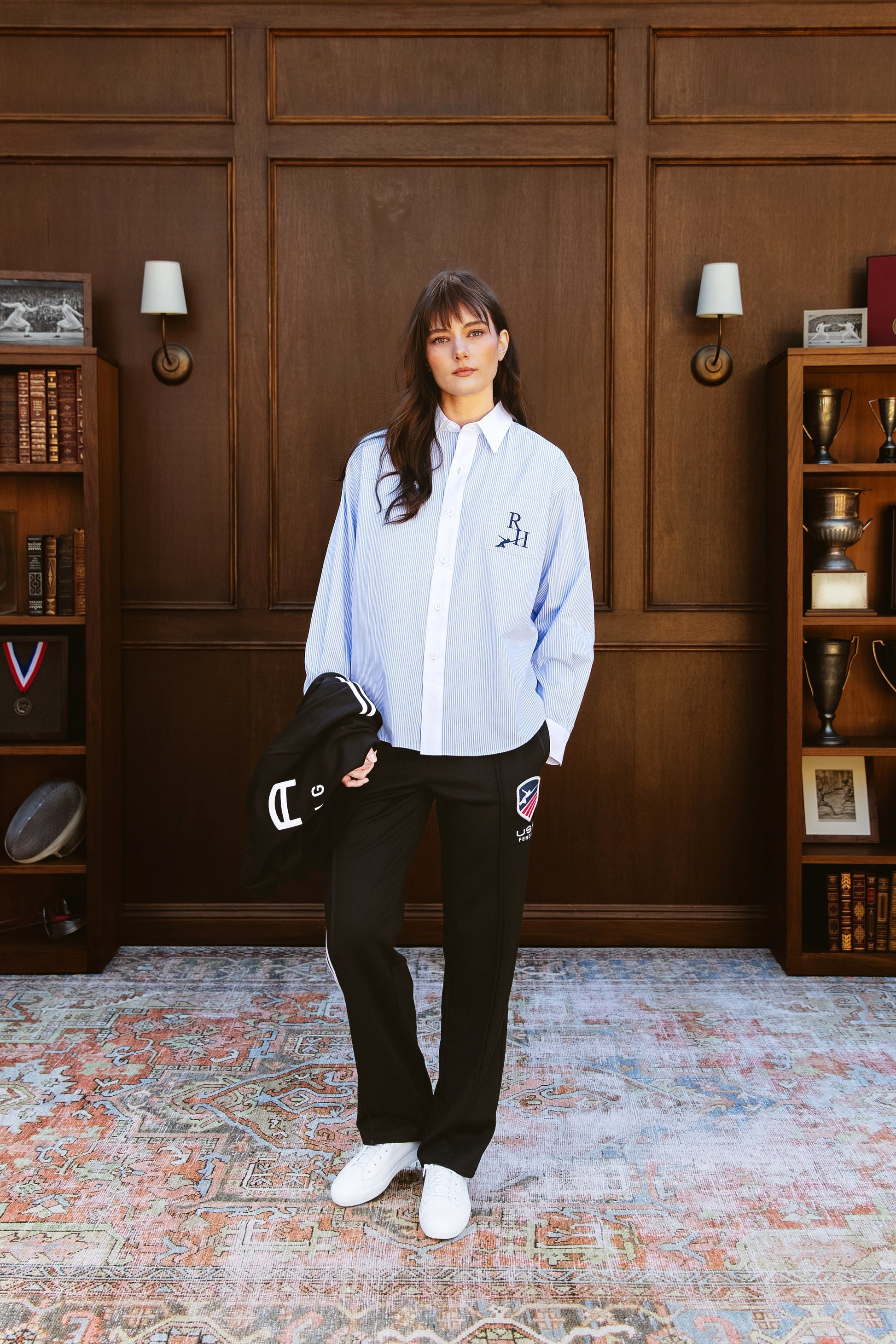 Woman in a light blue shirt and black pants standing in a room with wooden paneling and bookshelves.
