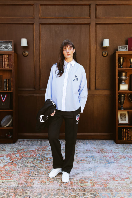 Woman in a light blue shirt and black pants standing in a room with wooden paneling and bookshelves.
