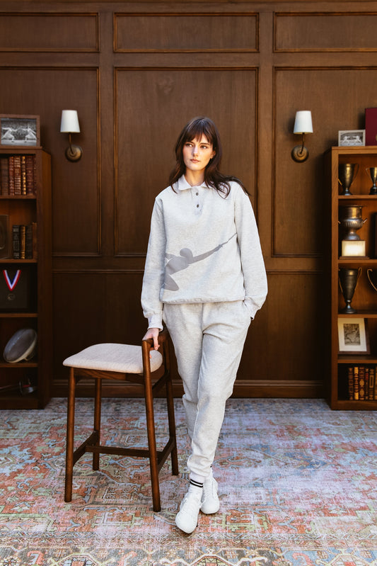 Woman in a light polo pullover with a fencing swordsman motif silhouette  on it in gray standing in a room with wooden paneling and bookshelves and other fencing or olympic memorabilia 